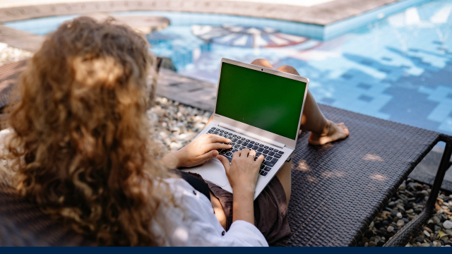 The image shows a female sitting with her back to the camera, an open laptop in front of her and appears to be working beside a swimming pool.