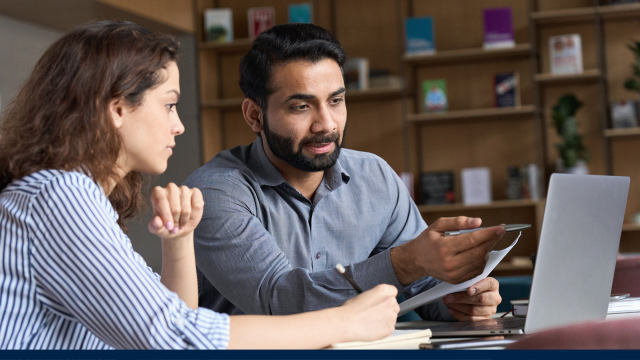 Image is of two people seated in front of an open laptop. It seems they are discussing something on the screen. One is talking while the other listens intently.