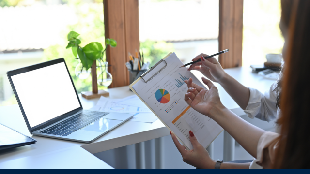 The image shows people in front of an open laptop discussing data on a clipboard held up in front of them.