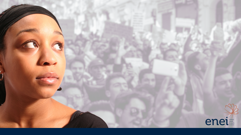 The background is a black and white semi-transparent image of many people involved in a protest, holding placards and shouting aggressively. Set against this, in full colour, is a Black female looking off into the distance and looking anxious and pensive, as if she is viewing the scene in her head.