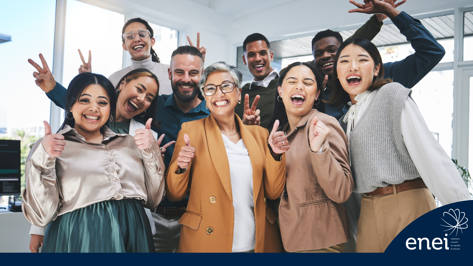 A diverse group of colleagues smiling, some giving thumbs up and peace signs, in a well-lit office environment.